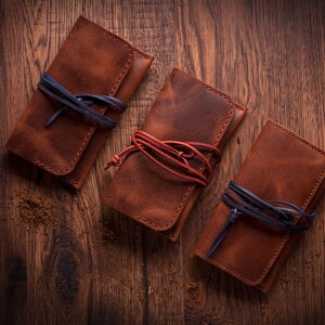 May include: Three brown leather pouches with a flap closure and a tie closure. The pouches are tied with blue and red cords. The pouches are on a wooden surface.