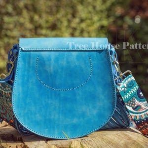 Op de afbeelding: Een blauwe leren schoudertas met een klep sluiting en een gepatroneerde band. De tas heeft een franje detail en een gestikt ontwerp aan de voorkant. De band is gemaakt van stof met een paisley patroon.