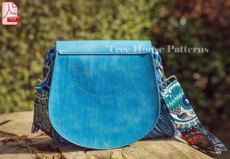 Op de afbeelding: Een blauwe leren schoudertas met een klep sluiting en een gepatroneerde band. De tas heeft een franje detail en een gestikt ontwerp aan de voorkant. De band is gemaakt van stof met een paisley patroon.