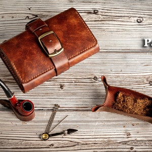 May include: A brown leather tobacco pouch with a buckle closure, a red pipe, a small metal tool, and a brown leather tobacco tray filled with tobacco.