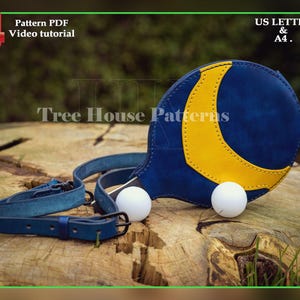Peut inclure: Un sac en cuir bleu et jaune avec un motif de croissant de lune, accompagné de deux balles blanches et d'une sangle en cuir bleu. Le sac est posé sur une surface en bois. Le texte sur l'image indique "Tree House Patterns".