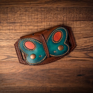 May include: A brown leather wallet with a butterfly design. The butterfly has teal wings with orange accents and brass rivets. The wallet is on a wooden surface.