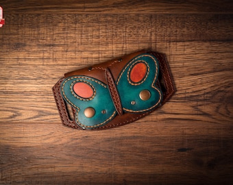 May include: A brown leather wallet with a butterfly design. The butterfly has teal wings with orange accents and brass rivets. The wallet is on a wooden surface.