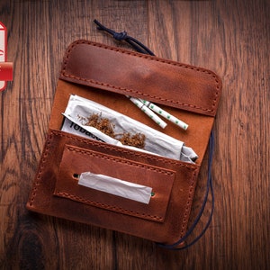 May include: A brown leather tobacco pouch with a flap that opens to reveal tobacco, rolling papers, and filters. The pouch has a blue cord and is on a wooden surface.