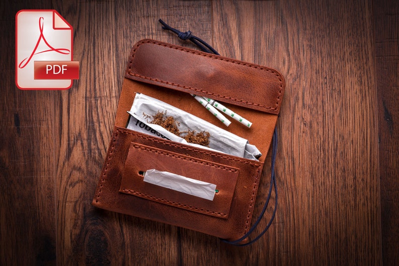 May include: A brown leather tobacco pouch with a flap that opens to reveal tobacco, rolling papers, and filters. The pouch has a blue cord and is on a wooden surface.
