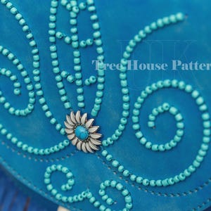 Op de afbeelding: Een turquoise blauwe leren tas met een decoratief ontwerp van turquoise kralen en een zilveren bloem met een turquoise centrum. De tas heeft een franje rand.