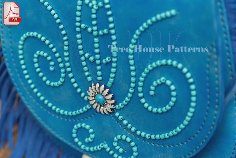 Op de afbeelding: Een turquoise blauwe leren tas met een decoratief ontwerp van turquoise kralen en een zilveren bloem met een turquoise centrum. De tas heeft een franje rand.