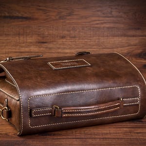 May include: A brown leather satchel bag with a top handle and a stitched design. The bag has a metal buckle and a label that says "Travel"