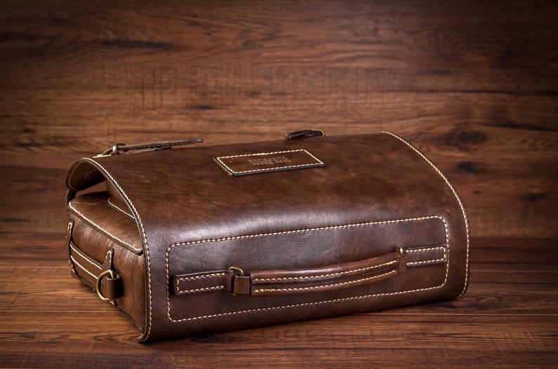 May include: A brown leather satchel bag with a top handle and a stitched design. The bag has a metal buckle and a label that says "Travel"