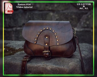 May include: A brown leather satchel bag with a flap closure and a buckle. The bag has a long strap and is decorated with metal studs. The bag is sitting on a rock.