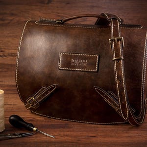 May include: A brown leather messenger bag with a long adjustable strap, a top handle, and a brass buckle. The bag has a stitched design and a label that reads "The Leather Satchel Co."