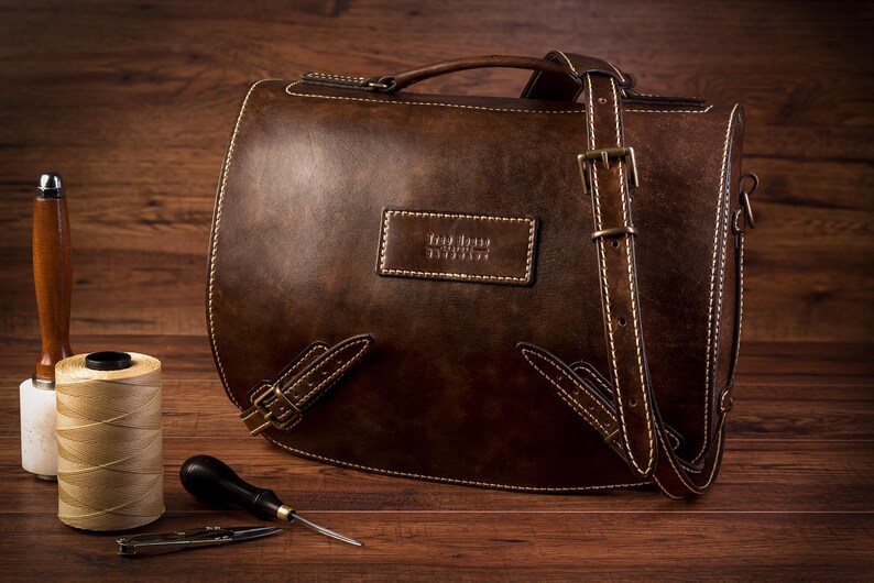 May include: A brown leather messenger bag with a long adjustable strap, a top handle, and a brass buckle. The bag has a stitched design and a label that reads "The Leather Satchel Co."