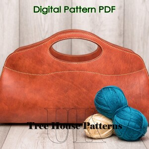 May include: A brown leather handbag with a top handle and a stitched design. The bag is sitting on a wooden surface with two balls of yarn, one teal and one cream.