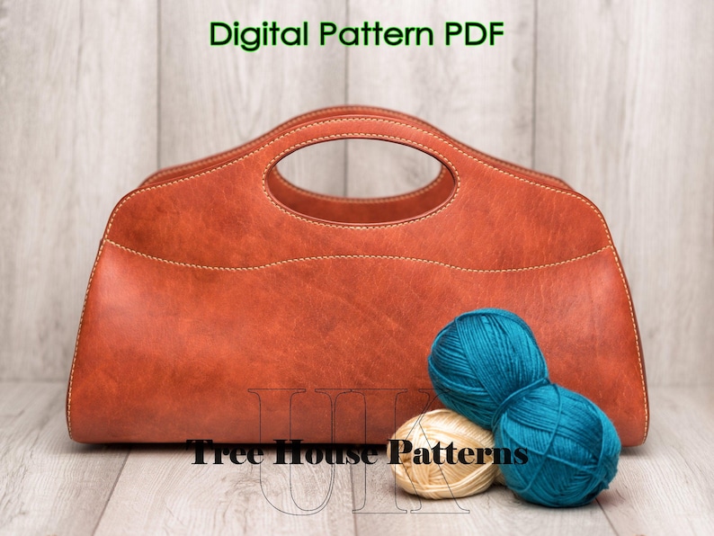 May include: A brown leather handbag with a top handle and a stitched design. The bag is sitting on a wooden surface with two balls of yarn, one teal and one cream.