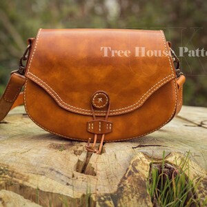 May include: A brown leather saddle bag with a flap closure and a strap. The bag has a white stitching detail and a metal buckle. The bag is sitting on a wooden surface.
