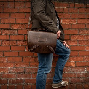 May include: A brown leather messenger bag with a flap closure and adjustable strap. The bag has a distressed look and is being worn by a person standing in front of a brick wall.
