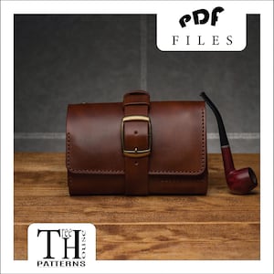 以下が含まれることがあります： 木製の表面に、真鍮のバックル留め具が付いた茶色の革製のタバコポーチと、茶色のパイプが置かれています。ポーチにはステッチのデザインが施され、パイプは茶色の木製のステムと黒いボウルを持っています。