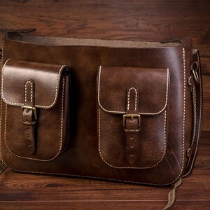 May include: A brown leather messenger bag with two front pockets and a long strap. The pockets have buckles and are stitched with white thread.