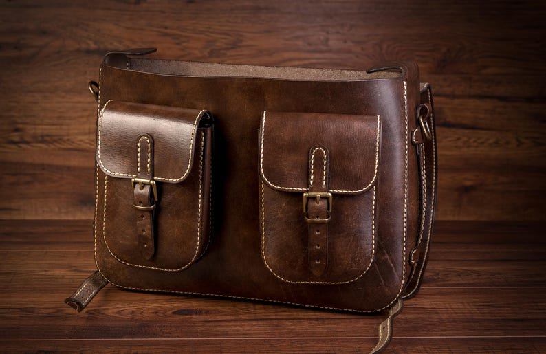 May include: A brown leather messenger bag with two front pockets and a long strap. The pockets have buckles and are stitched with white thread.