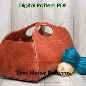May include: A brown leather tote bag with a stitched design and a handle. The bag is sitting on a wooden surface with balls of yarn in blue and white.