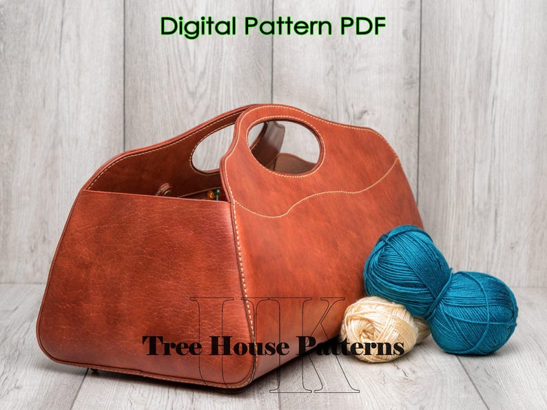 May include: A brown leather tote bag with a stitched design and a handle. The bag is sitting on a wooden surface with balls of yarn in blue and white.