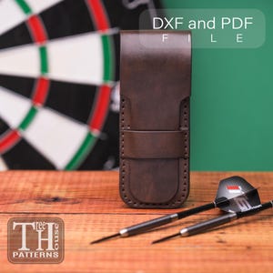 Puede incluir: Un estuche de dardos de cuero marrón con cierre de correa, junto a dos dardos con cañas negras y plumas estampadas. El texto "DXF and PDF FILE" es visible. El fondo incluye un tablero de dardos y una superficie de madera.