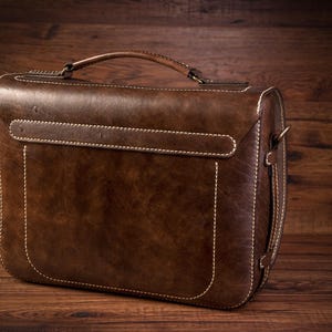 May include: A brown leather briefcase with a top handle and a strap. The briefcase has white stitching and a flap closure.