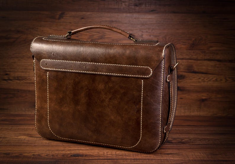 May include: A brown leather briefcase with a top handle and a strap. The briefcase has white stitching and a flap closure.