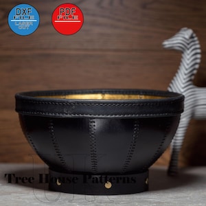 May include: A black leather bowl with gold accents and stitching. The bowl has a round base and is sitting on a wooden surface. The text "Tree House Patterns" is visible on the base of the bowl.