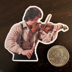 May include: A sticker featuring a man playing a violin. The man is wearing a light pink shirt, black suspenders, and black pants. A quarter is next to the sticker for size comparison. The violin is brown.