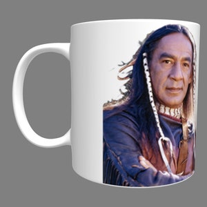 May include: White ceramic mug with a color image of a Native American man with long dark hair and a beaded headband. He is wearing a blue and brown fringed jacket and has a serious expression.