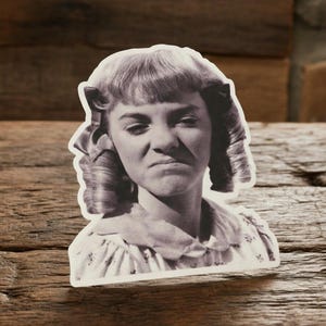 May include: A monochrome sticker showing a vintage image of a woman with a displeased expression. The woman has curly hair and is wearing a collared dress. The sticker is die-cut with a white border.