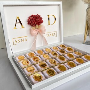 Może przedstawiać: Białe pudełko wypełnione czekoladkami, każda zawinięta w różową i złotą folię. Wieko pudełka zawiera złote litery i małą kompozycję kwiatową z różową kokardą. Tekst na pokrywie brzmi "A & D" i "ANNA & DARL".