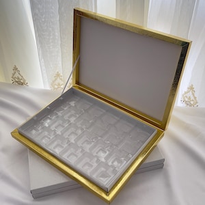 Könnte beinhalten: Eine offene Schmuckschatulle mit goldfarbenem Rahmen und weißem Innenraum. Die Box hat einen transparenten Kunststoffeinsatz mit mehreren Fächern. Der Deckel ist ebenfalls weiß mit einem goldenen Rand. Die Box steht auf einer weißen Box.