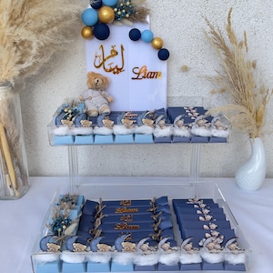 Può includere: Un'esposizione a due livelli di cioccolatini, decorata per una festa. I cioccolatini sono avvolti in blu navy e azzurro, con motivi di orsetti e nuvole. Un cartello con scritte dorate e un orsetto è posto sopra l'esposizione.