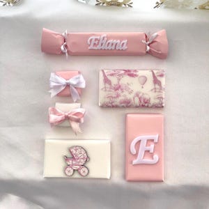 Könnte beinhalten: Eine Sammlung von rosa und weiß verpackten Süßigkeiten und Schokoladen. Eine Süßigkeit ist mit dem Namen "Eliana" verpackt. Andere Pralinen haben einen Kinderwagen, den Buchstaben "E" und eine Safari-Szene mit Giraffen und einem Elefanten.
