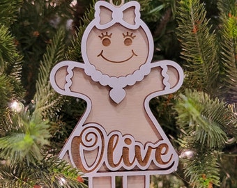 May include: A wooden Christmas ornament in the shape of a gingerbread person, with the name "Olive" cut out. The ornament has a light brown base with white accents and is hanging on a Christmas tree.