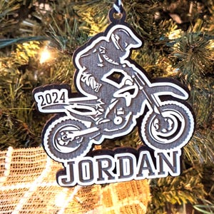 May include: A wooden Christmas bauble depicting a silhouette of a motocross rider on a dirt bike. The bauble is painted in shades of grey and black and includes the year "2024" and the name "JORDAN".