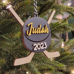 以下が含まれることがあります： 黒いホッケーのパックのオーナメントで、金色の「Judah 2023」の文字と、木製のホッケーのスティックが2本付いている