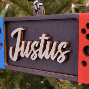 Puede incluir: Un adorno navideño de madera con forma de consola Nintendo Switch con un mando azul y rojo. El adorno está personalizado con el nombre "Justus" en letras blancas.