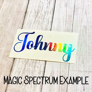 May include: A close-up of a white sticker with the name "Johnny" written in a rainbow holographic font. The sticker is on a light wood surface. The text "MAGIC SPECTRUM EXAMPLE" is written below the sticker.