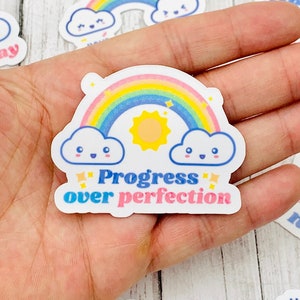 May include: A set of rainbow-themed stickers with positive messages. The stickers feature rainbows, clouds, and stars. The text on the stickers includes "Progress over perfection", "Healing not linear", "Okay", and "Mental Health".