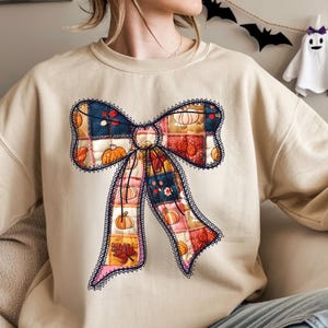 May include: A beige sweatshirt featuring a large bow design. The bow is made of patchwork squares with pumpkin and floral patterns in orange, red, and blue. The bow is outlined with a dark blue lace trim.