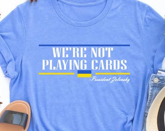 May include: A light blue t-shirt with the text "We're not playing cards - President Zelensky" in white. The Ukrainian flag is displayed in yellow and blue.