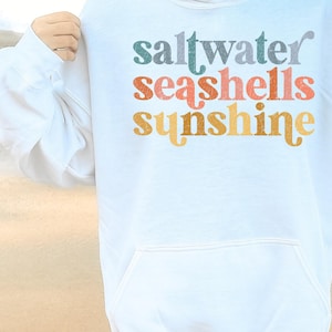 May include: White hoodie with a graphic print that says "saltwater seashells sunshine" in colorful retro lettering.