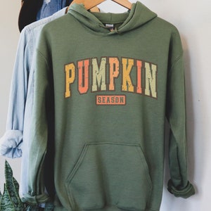 Może przedstawiać: Zielona bluza z kapturem z napisem "PUMPKIN SEASON" w stylu vintage, w kolorze pomarańczowym, ż&oacute;łtym i zielonym.