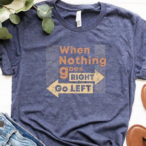 May include: Navy blue t-shirt with the text "When Nothing goes RIGHT Go LEFT" in orange and beige. The design includes an arrow pointing left. The shirt is made of a soft material.