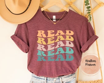 Könnte beinhalten: Ein maroonfarbenes T-Shirt mit dem Wort "READ", das fünfmal in einem Regenbogen aus Farben wiederholt wird. Das Shirt hängt an einem Kleiderbügel und hat eine lockere Passform.