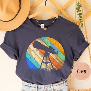 May include: A navy blue t-shirt with a retro-style graphic of a telescope in front of a colorful striped circle. The shirt is on a hanger and has a brown hat and a straw bag in the background.