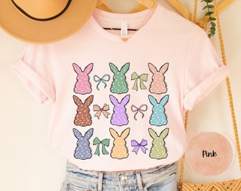 May include: Pink t-shirt with a pattern of colorful bunnies with polka dots and bows. The shirt is on a hanger and has a light pink background.